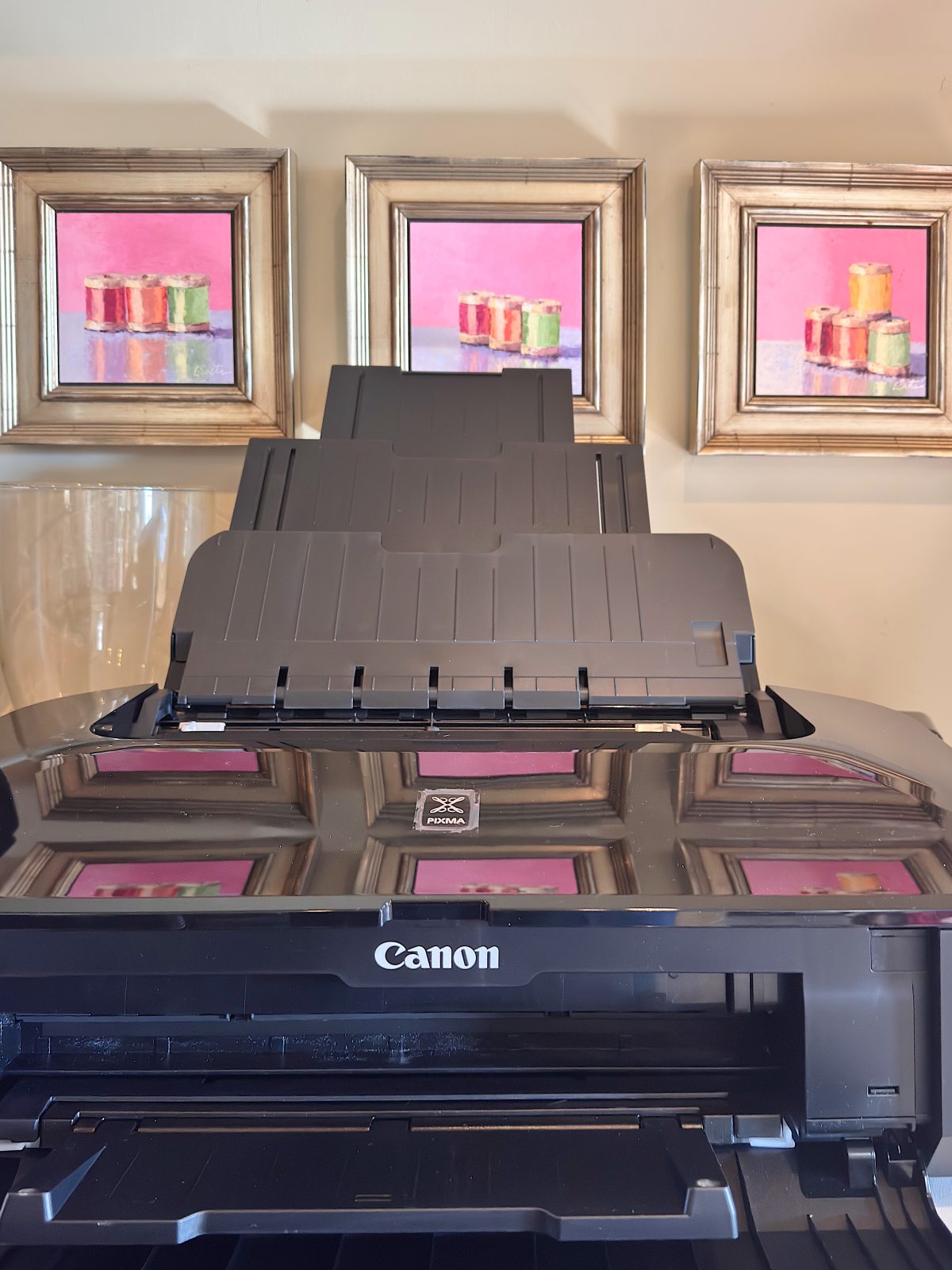 A Canon printer sits in front of a wall with three framed pink paintings featuring colorful cakes or desserts.
