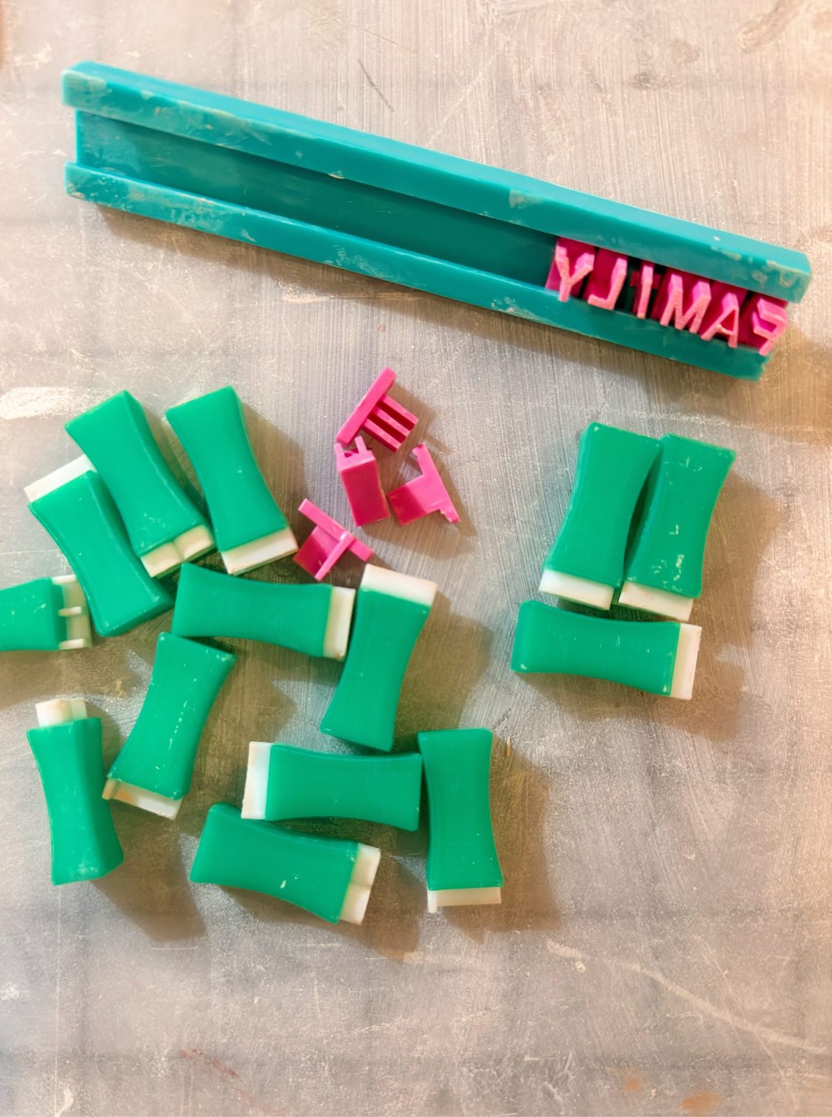 Green and white clay pieces are scattered on a surface, with pink plastic letter stamps and teal handles positioned above them.