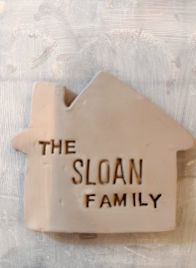 A beige ceramic house-shaped plaque with the words "THE SLOAN FAMILY" engraved on the front.