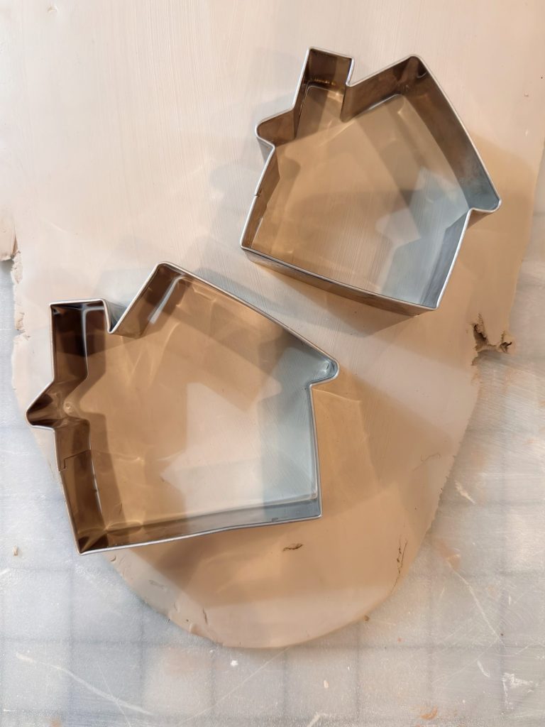 Two metal cookie cutters in the shape of houses placed on a light-colored, textured surface.