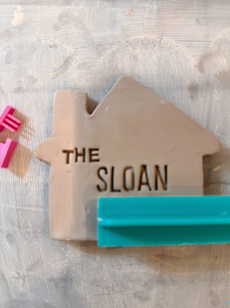 A gray clay house shape with the words "THE SLOAN" stamped on it, resting on a light surface with a blue tool and pink letter stamps nearby.