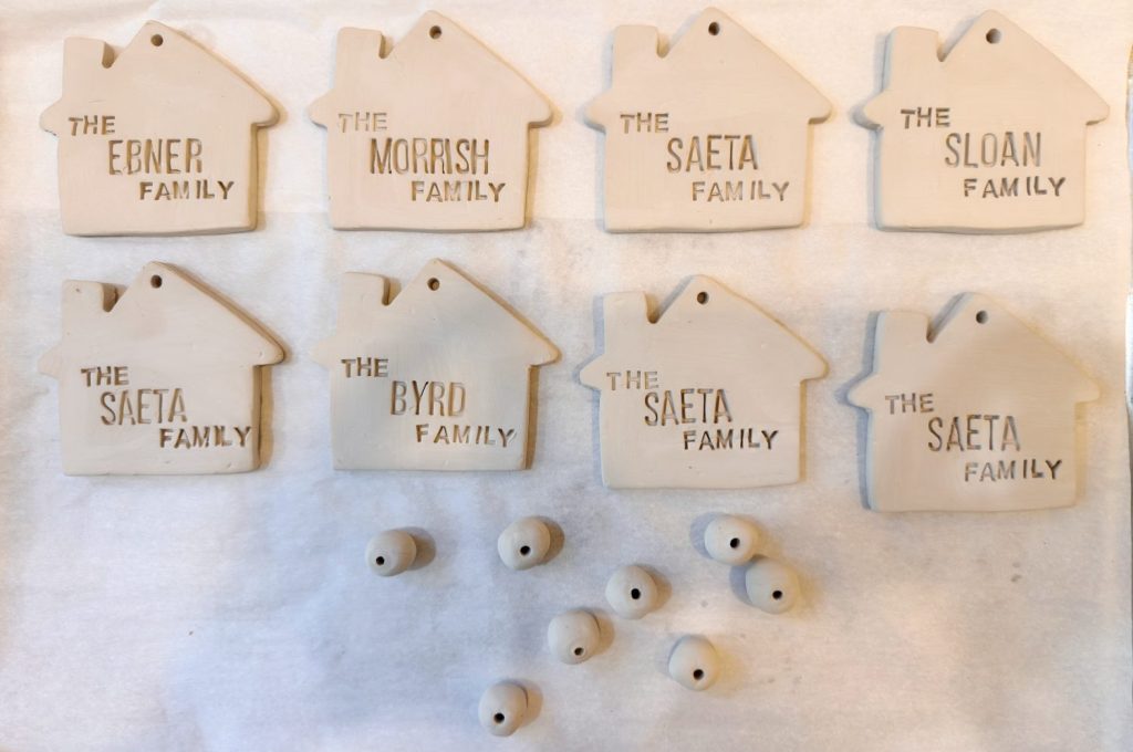 Seven unpainted, house-shaped clay ornaments labeled with different family names and several small clay beads arranged on a white surface.