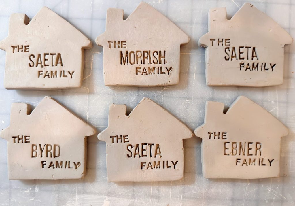 Six clay plaques shaped like houses, each labeled with different family names: SAETA, MORRISH, BYRD, and EBNER, and the words “THE FAMILY” beneath each name.