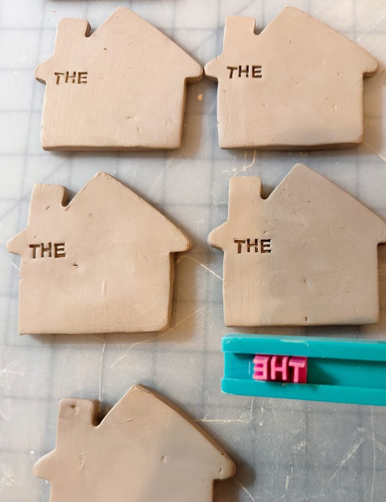 Five clay house shapes, four stamped with the word "THE," sit on a surface. A blue tool with pink letter stamps spelling "THE" is visible in the foreground.