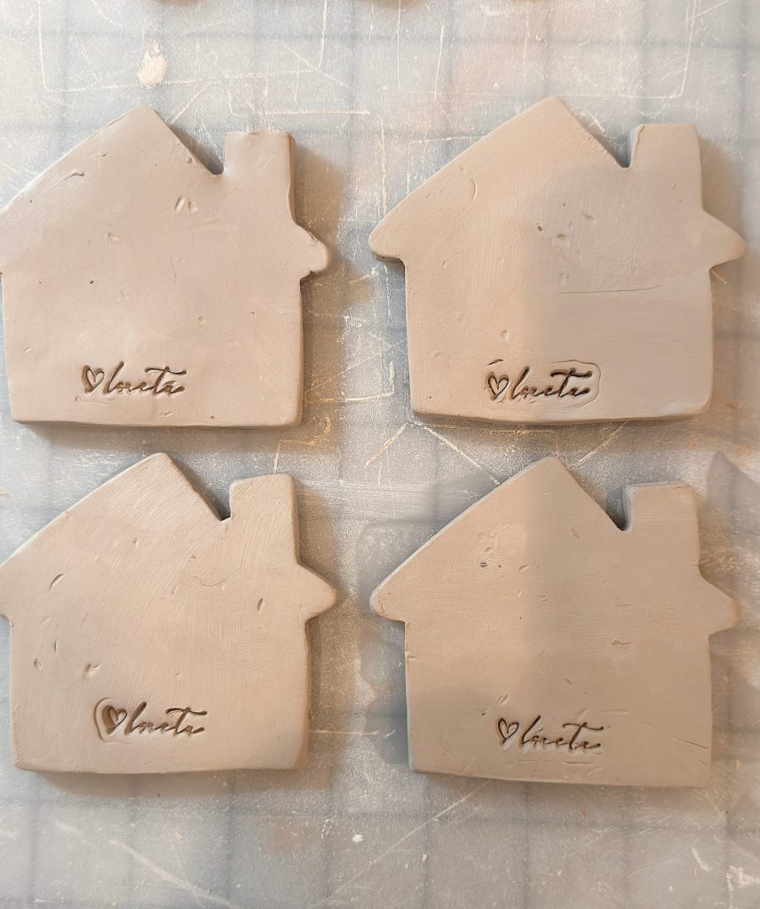 Four unglazed clay tiles shaped like houses, each stamped with the word "love" and a heart symbol, are arranged on a gridded work surface.