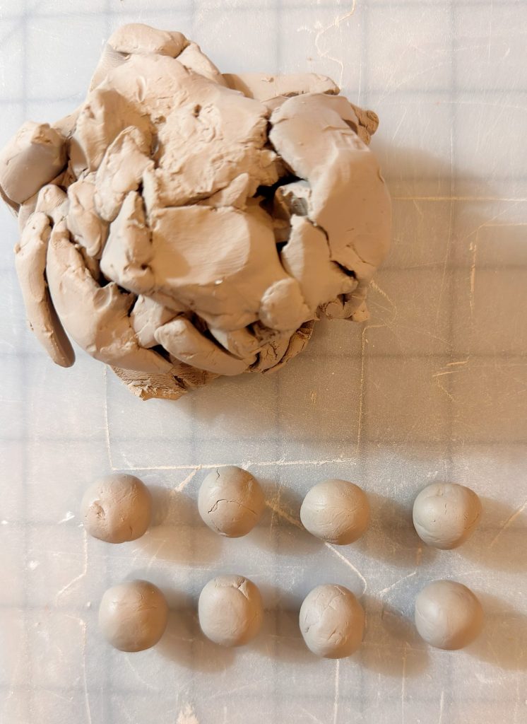 A large lump of clay sits above seven small clay balls arranged in a row on a gridded surface.
