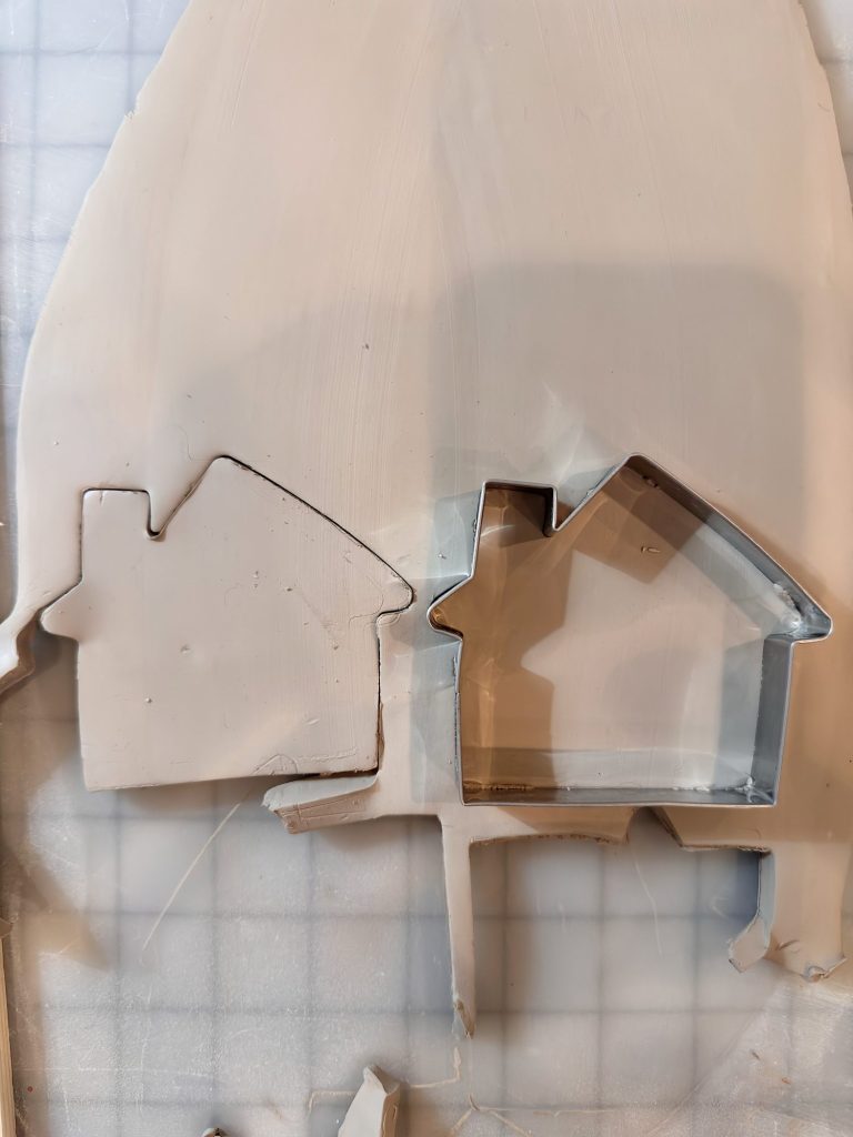 Metal house-shaped cookie cutter pressed into a sheet of clay, with one house shape already cut out beside the cutter.