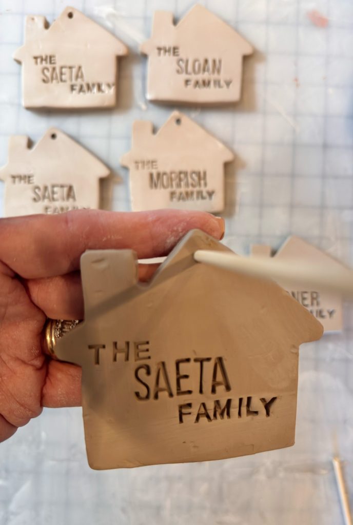 A hand holds a clay ornament shaped like a house with "THE SAETA FAMILY" stamped on it; similar ornaments labeled with different family names are in the background.