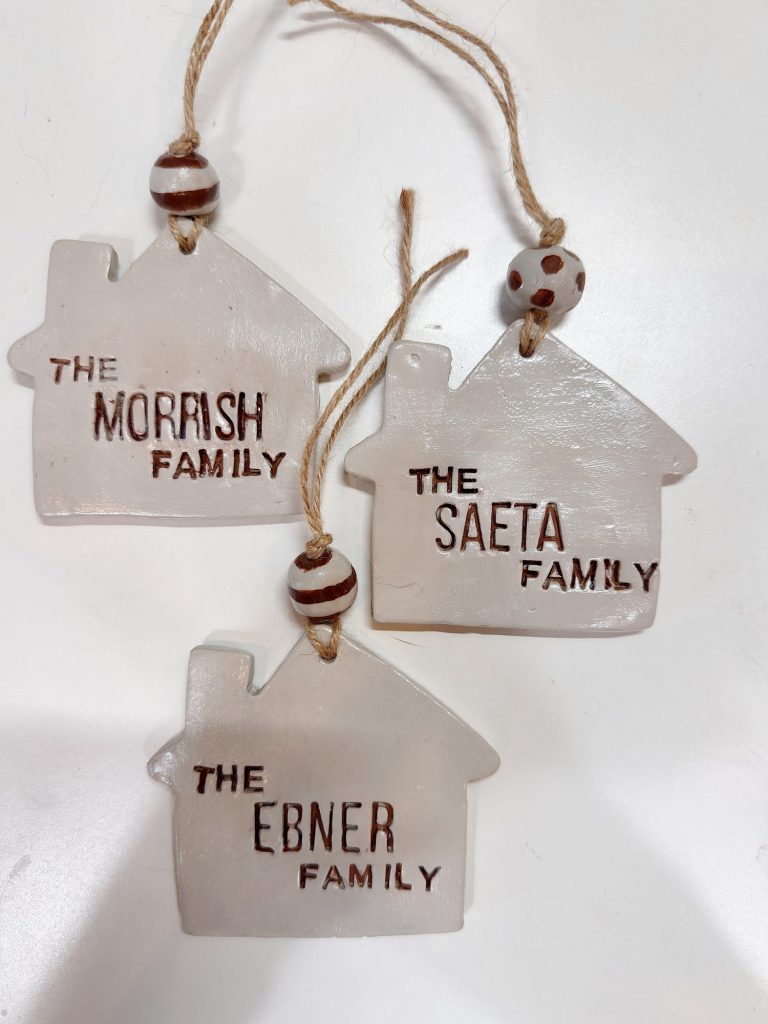 Three house-shaped ornaments with "The Morrish Family," "The Saeta Family," and "The Ebner Family" written on them, each hanging by a string with a decorative bead.