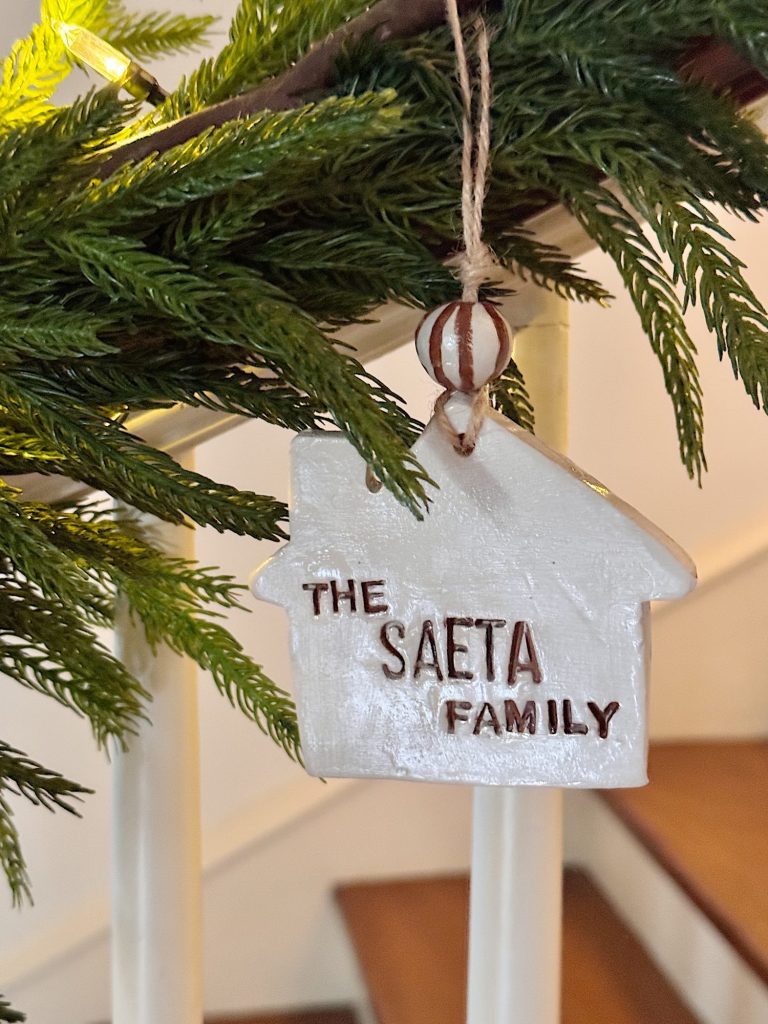 A white ceramic house ornament labeled "The Saeta Family" hangs from a railing decorated with green pine branches.