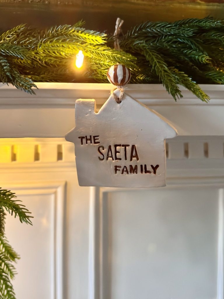 A white, house-shaped ornament labeled "The Saeta Family" hangs from a garland of greenery with a string light above it.