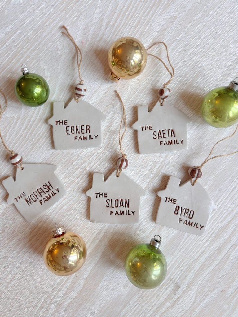 Four ceramic house-shaped ornaments labeled with family names are arranged on a white surface among gold and green Christmas baubles.