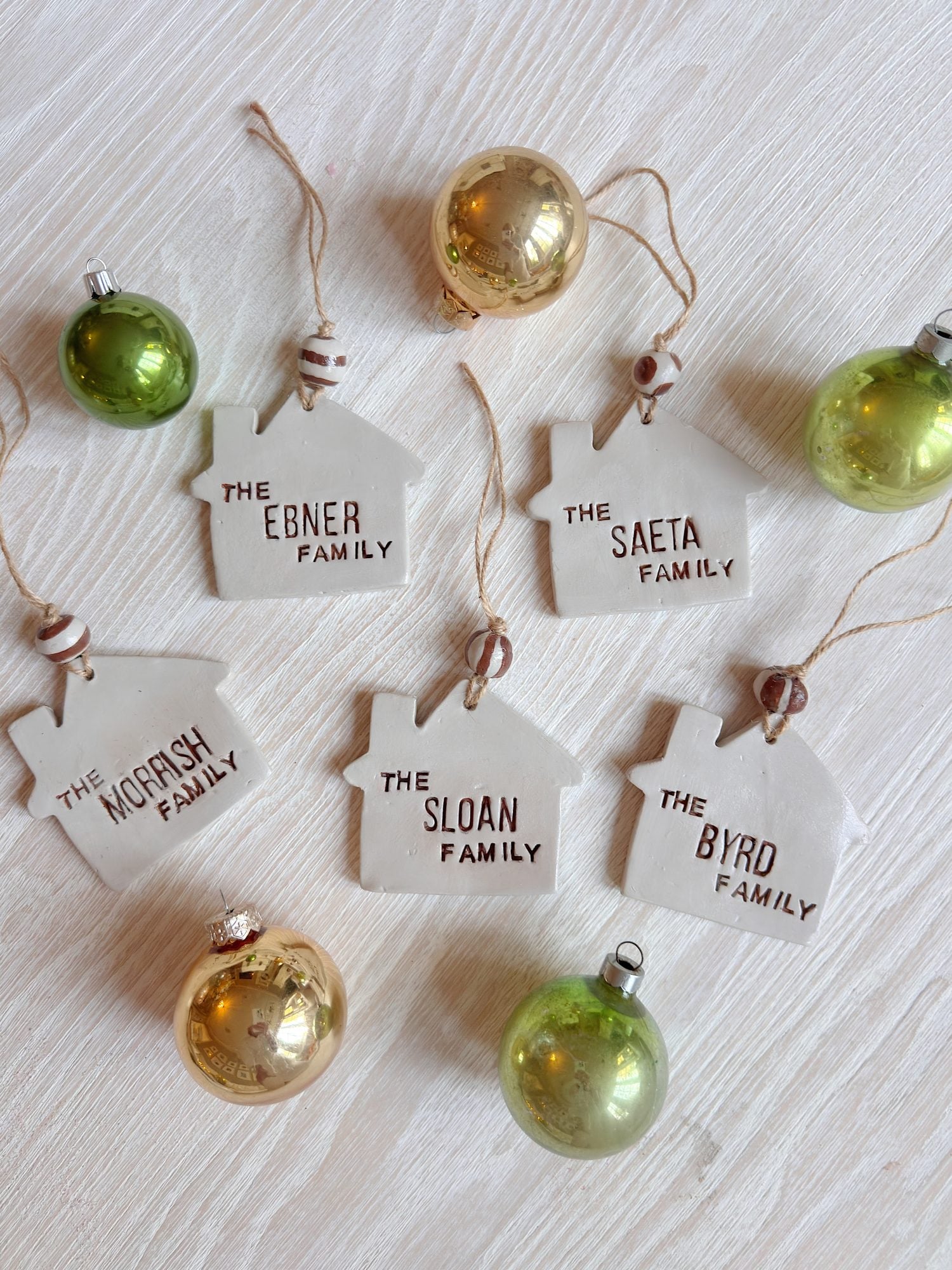 Four ceramic house-shaped ornaments labeled with family names are arranged on a white surface among gold and green Christmas baubles.