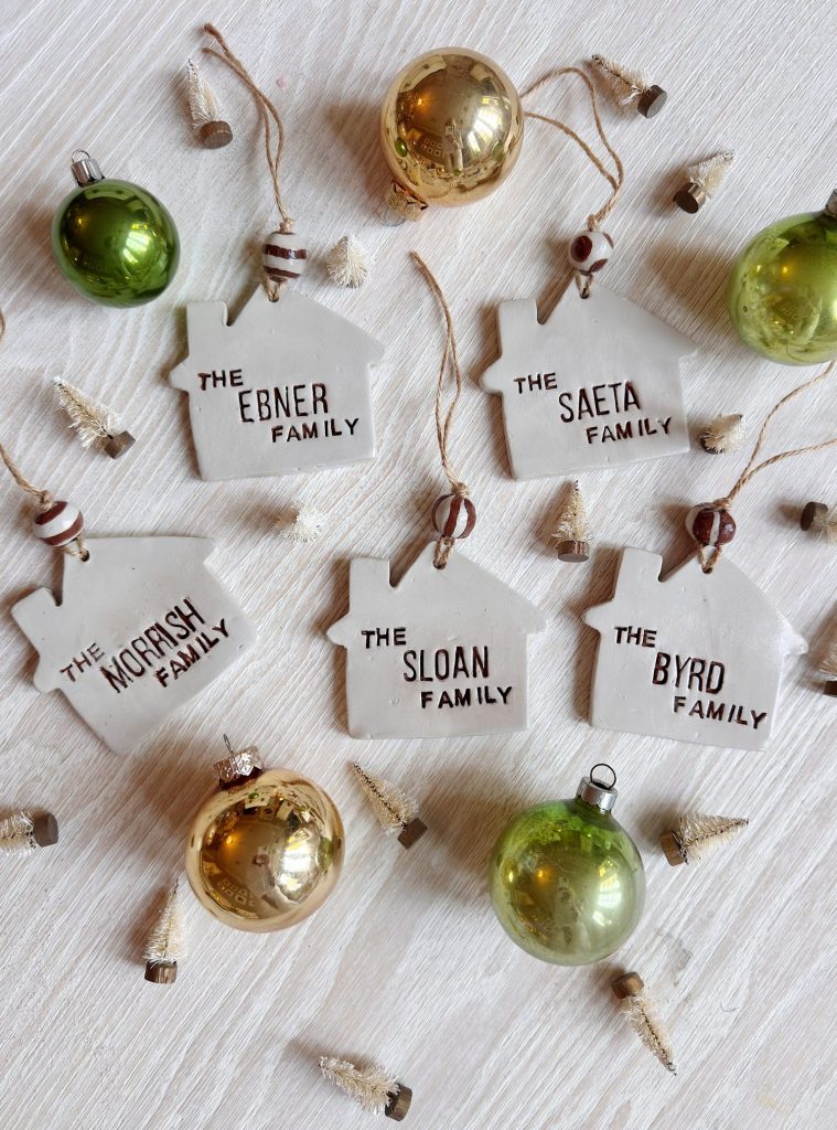 Five ceramic house-shaped ornaments labeled with different family names and surrounded by green and gold Christmas ornaments and miniature trees on a light wood surface.