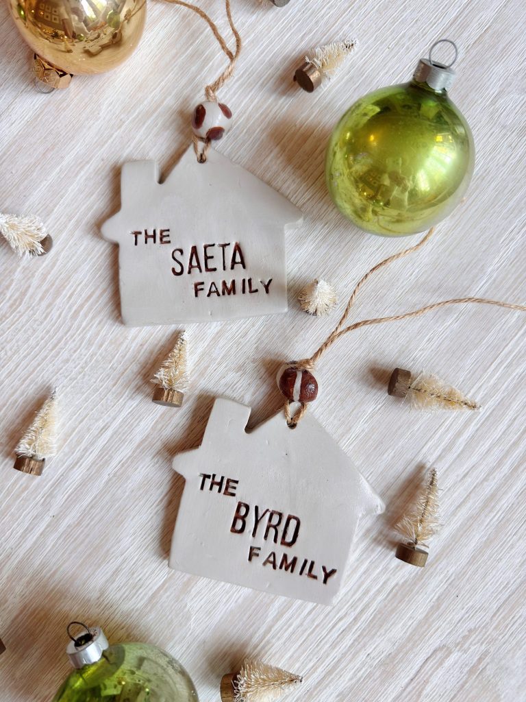Two house-shaped ornaments labeled "The Saeta Family" and "The Byrd Family" lie on a white surface with green ornaments and small decorative trees nearby.