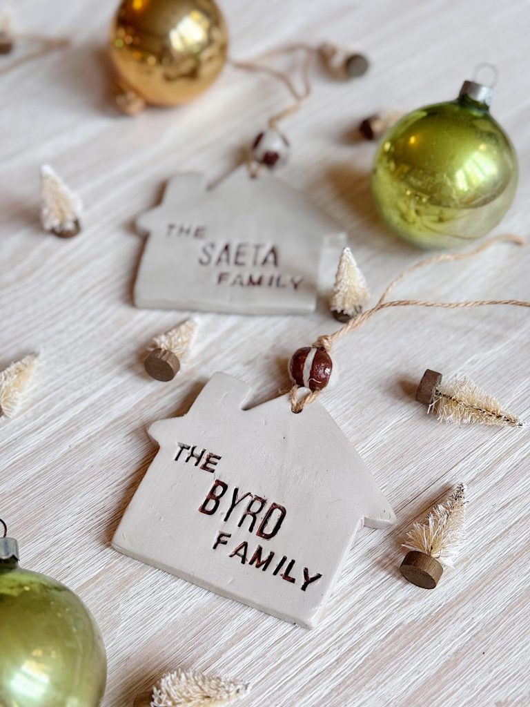 Two house-shaped clay ornaments labeled "The BYRD Family" and "The SAETA Family" lie on a wooden surface, surrounded by green and gold baubles and small decorative trees.