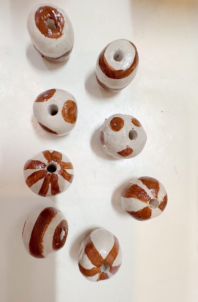 Eight round, clay beads with brown and white painted patterns are arranged on a white surface. Each bead has a central hole for stringing.
