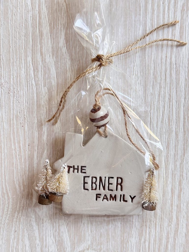 A ceramic house-shaped ornament labeled "The Ebner Family," decorated with two small model trees, wrapped in clear plastic with twine.