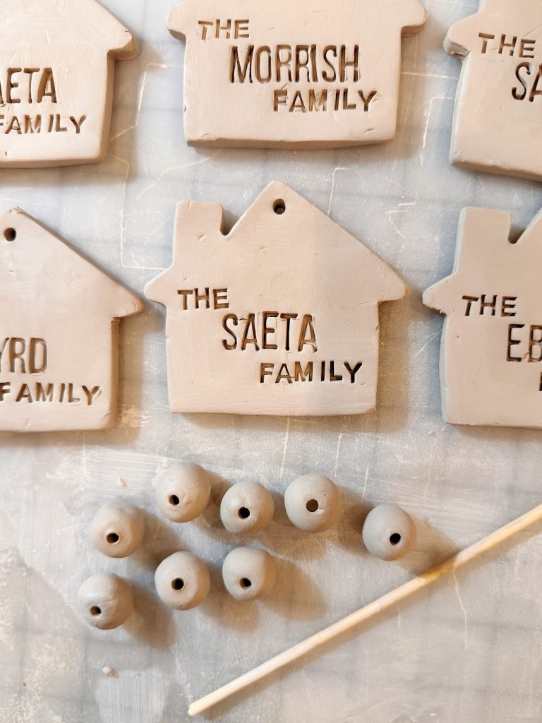 Clay house-shaped tags with family names, round clay beads, and a wooden stick arranged on a textured surface.
