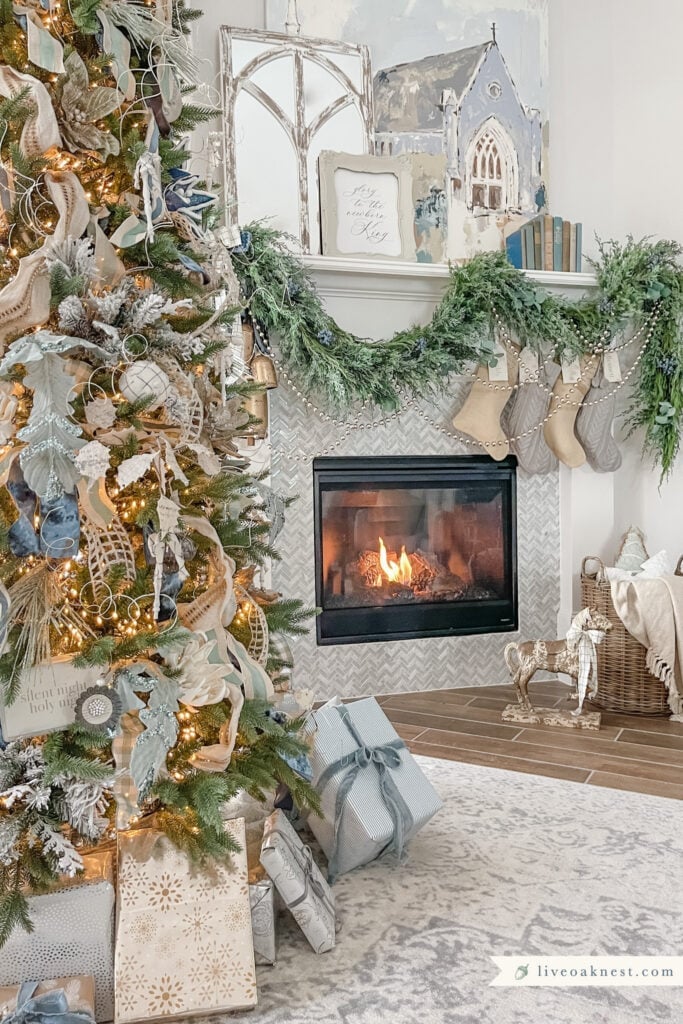 A decorated Christmas tree with wrapped presents stands near a lit fireplace, above which are stockings, greenery, and holiday decor in a cozy living room.