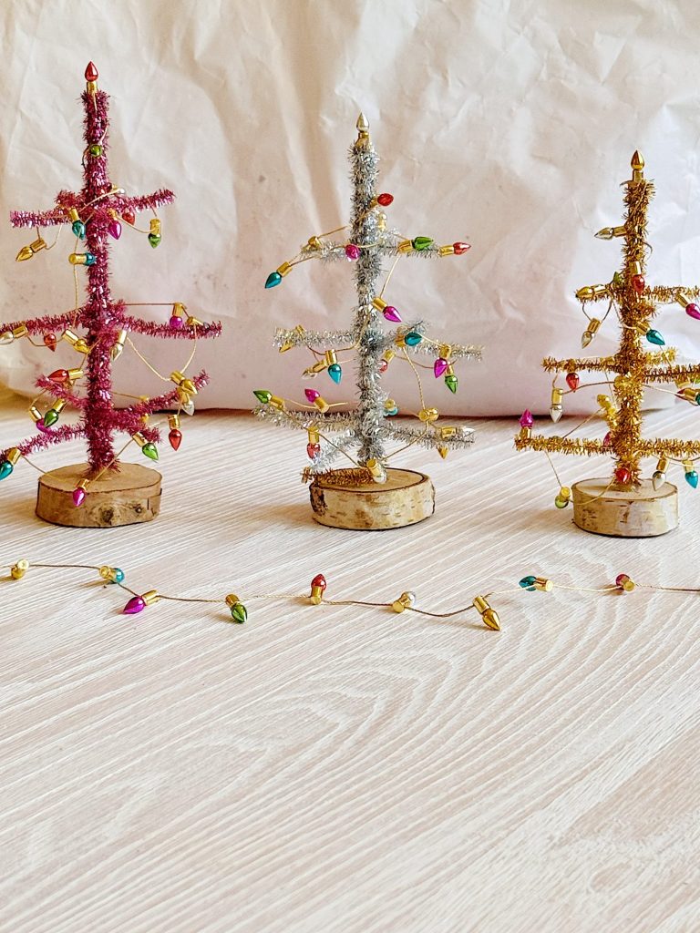 Three small decorative Christmas trees made of tinsel in red, silver, and gold stand on wooden bases, adorned with tiny colorful lights on a light wooden surface.