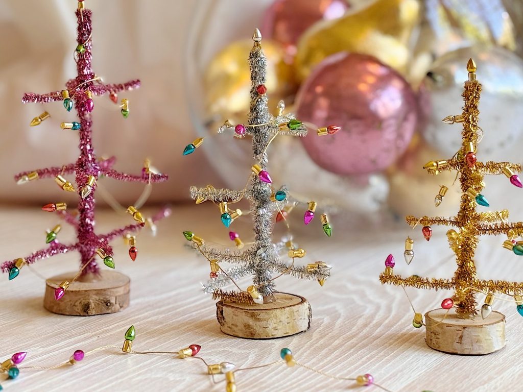Three small Christmas trees made of tinsel and wood sit on a light surface, decorated with colorful mini lights; blurred ornaments are in the background.