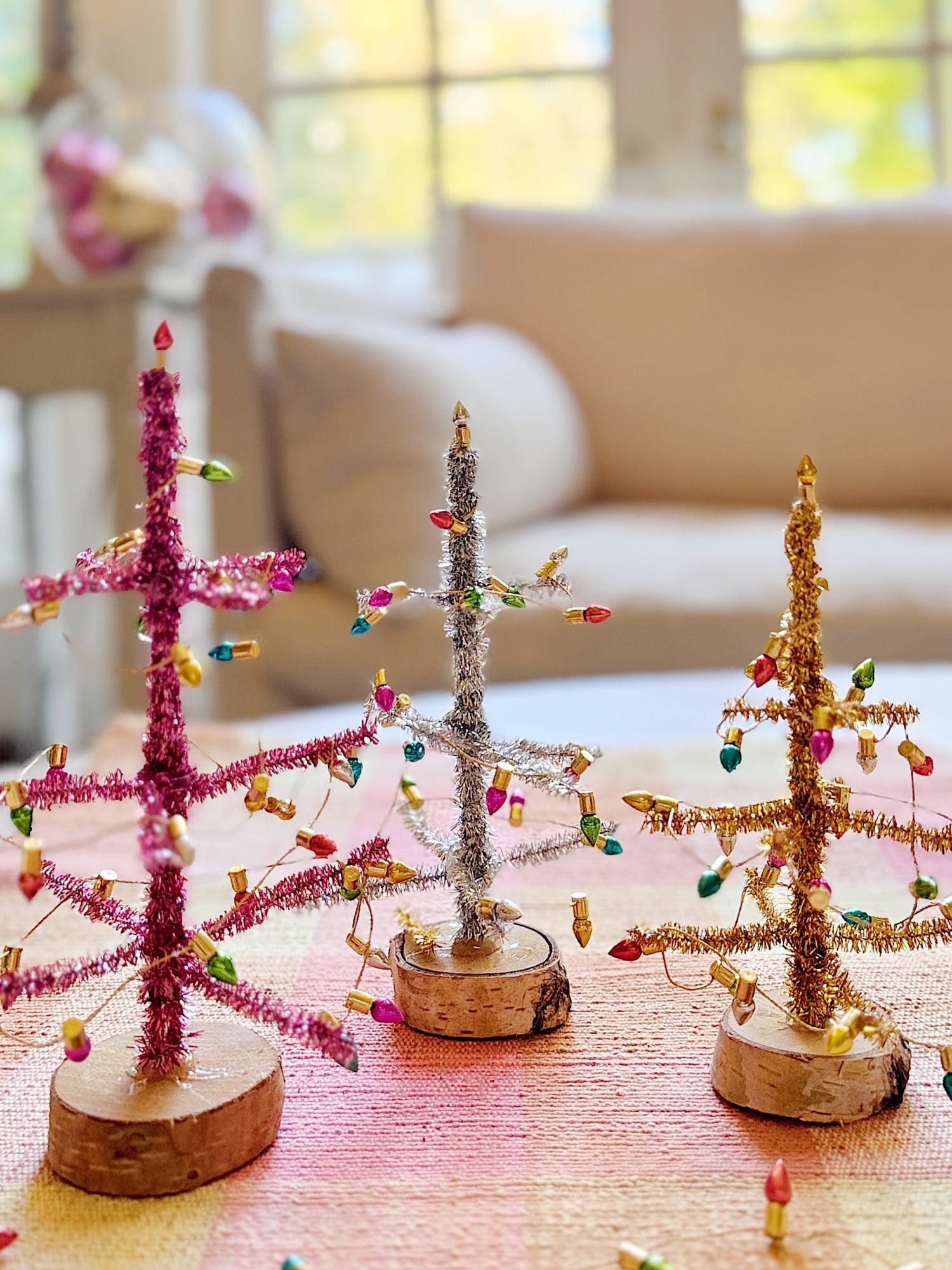 Three small tabletop Christmas trees made of colored tinsel and mini lights sit on a wooden table in a brightly lit living room.