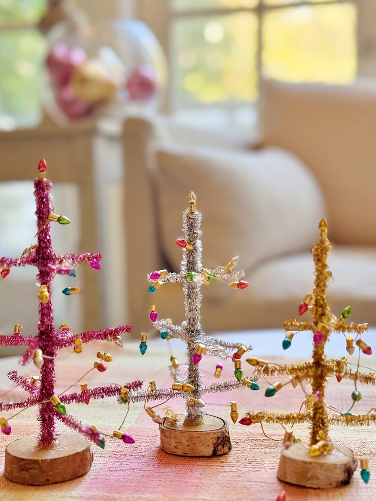 Three small, colorful, tinsel Christmas trees with wooden bases are displayed on a table in a brightly lit room with a sofa and window in the background.