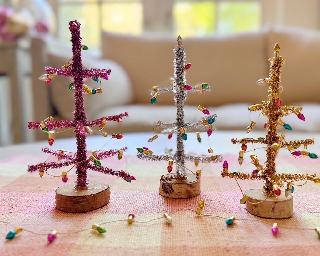 Three small decorative Christmas trees made of colored tinsel and string lights stand on a table with a plaid cloth, with a soft-focus couch and window in the background.