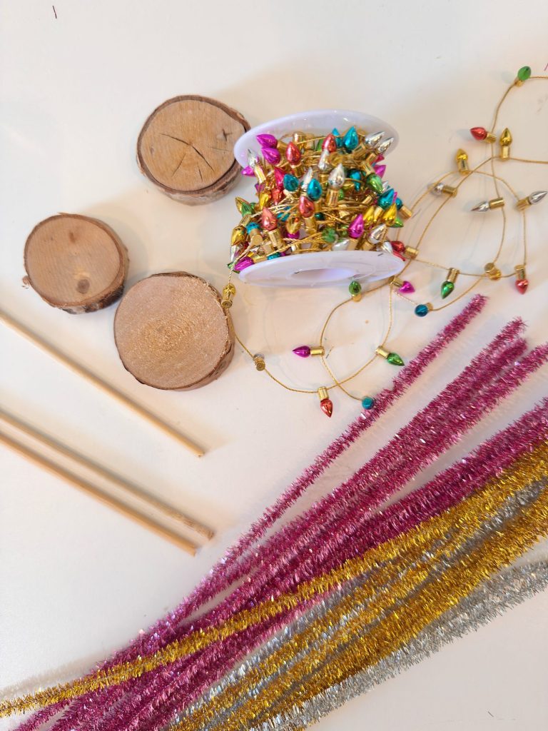 Wood slices, wooden dowels, colorful mini string lights, and metallic gold, pink, and silver pipe cleaners arranged on a white surface.