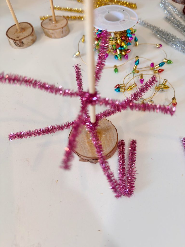 A craft project with pink pipe cleaners attached to a wooden base, surrounded by decorative materials including colorful string lights and a spool on a white surface.