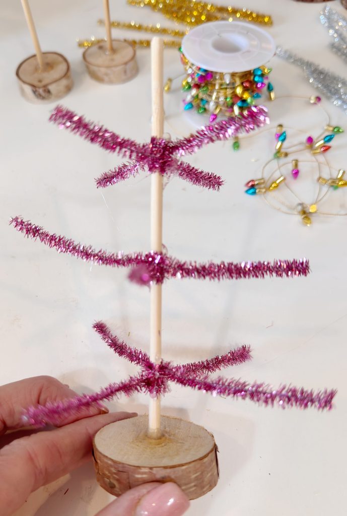 A hand holds a small DIY Christmas tree made from pink pipe cleaners on a wooden base, with craft supplies scattered in the background.