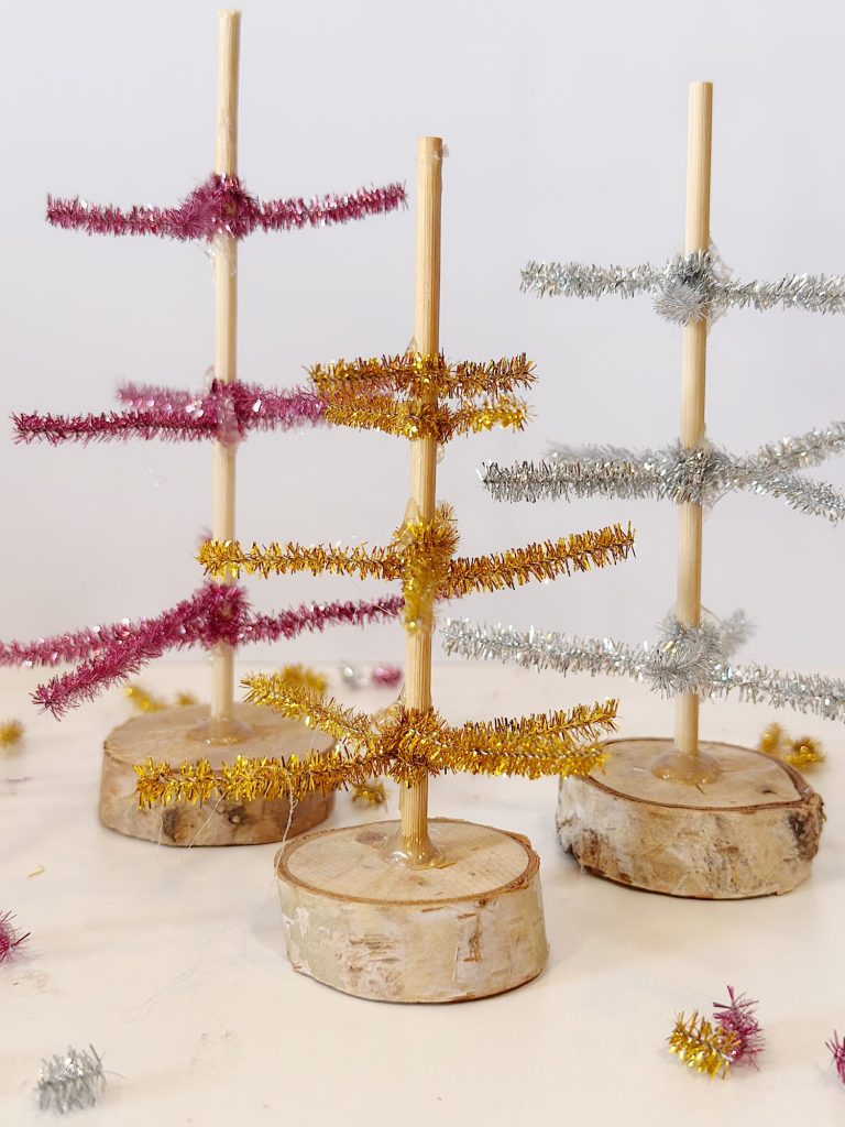Three small DIY Christmas trees made of wooden sticks, wood slices as bases, and metallic pipe cleaners in gold, pink, and silver arranged on a light surface.