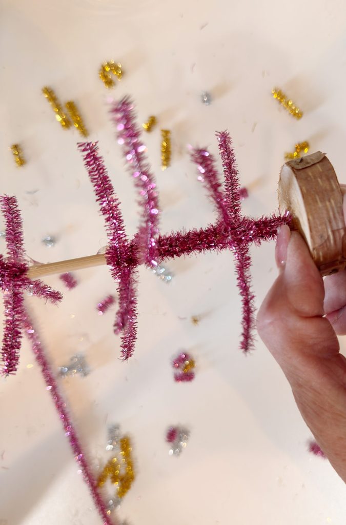 A hand holds a wooden base with pink pipe cleaners arranged in tiers on a stick; gold and silver pipe cleaners are scattered on a white surface.
