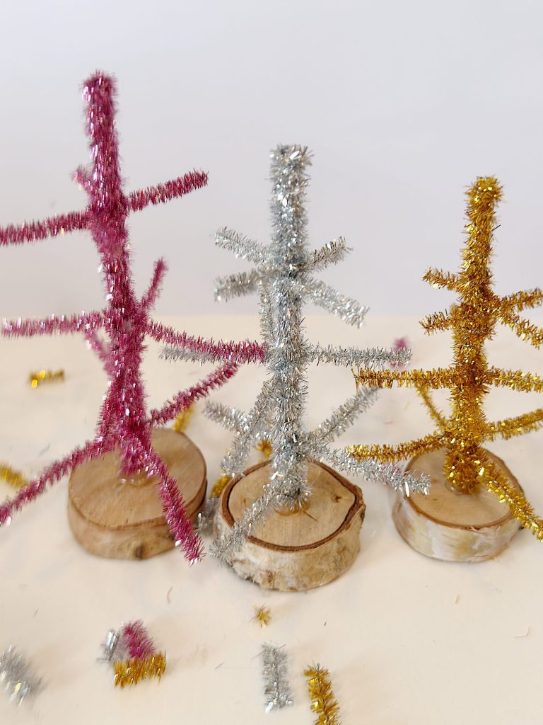Three small, handmade trees made of pink, silver, and gold tinsel pipe cleaners stand on round wooden bases against a plain background.