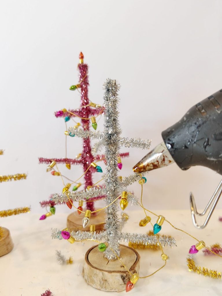 A hot glue gun is used to attach mini Christmas lights to a small, silver, pipe cleaner tree on a wooden base. Other decorated trees are in the background.