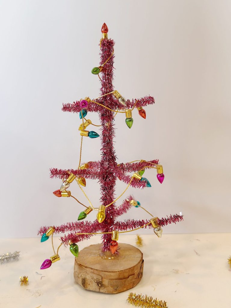 A small pink tinsel Christmas tree with colorful light bulb ornaments stands on a round wooden base against a plain light background.