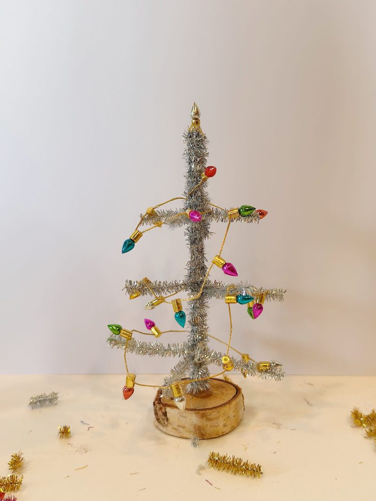 A small artificial Christmas tree made of silver tinsel stands on a wooden base, decorated with a string of colorful lights.