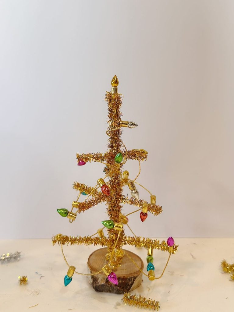 A small handmade Christmas tree made from gold tinsel and colorful bulb ornaments, standing on a wooden base against a plain background.