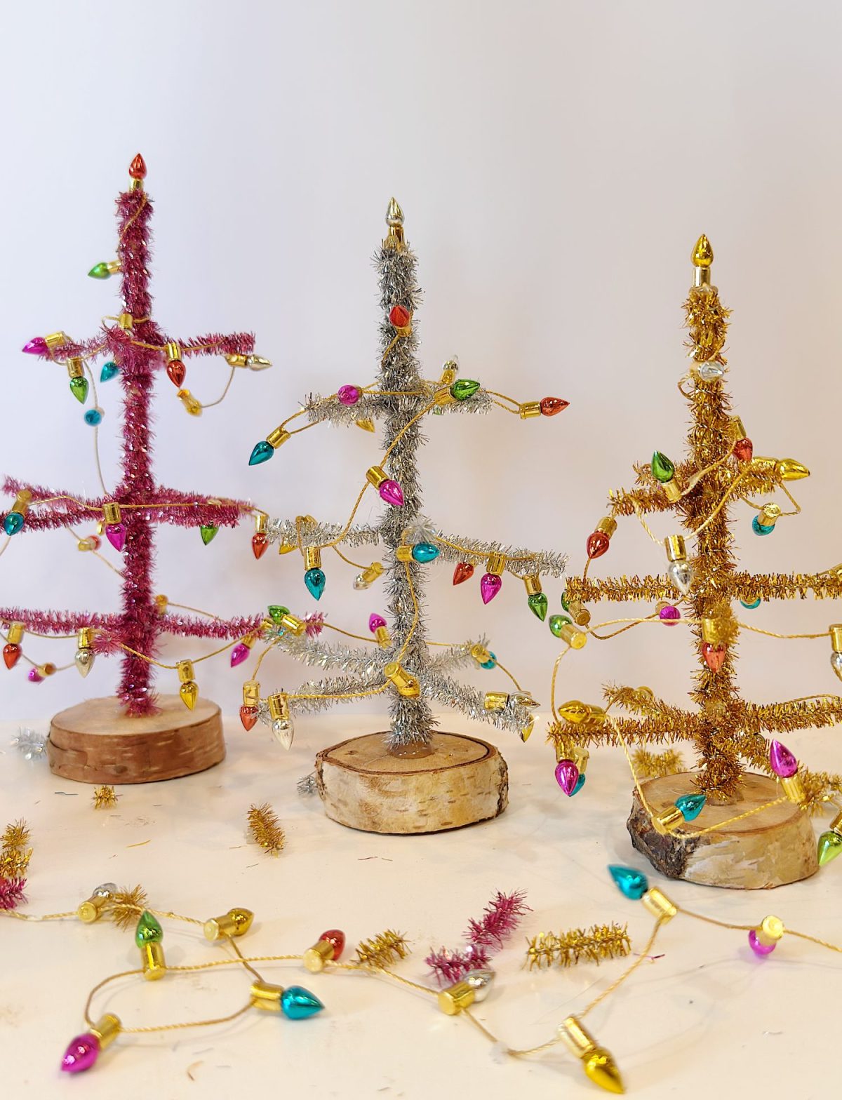 Three small tabletop Christmas trees made of tinsel in pink, silver, and gold, with wooden bases and miniature colorful string lights, set against a plain background.
