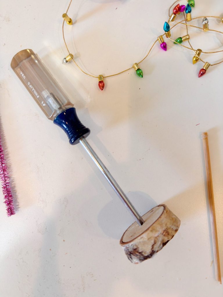 A flathead screwdriver is inserted into a round wooden disc, lying on a white surface near string lights, a pink pipe cleaner, and a wooden stick.
