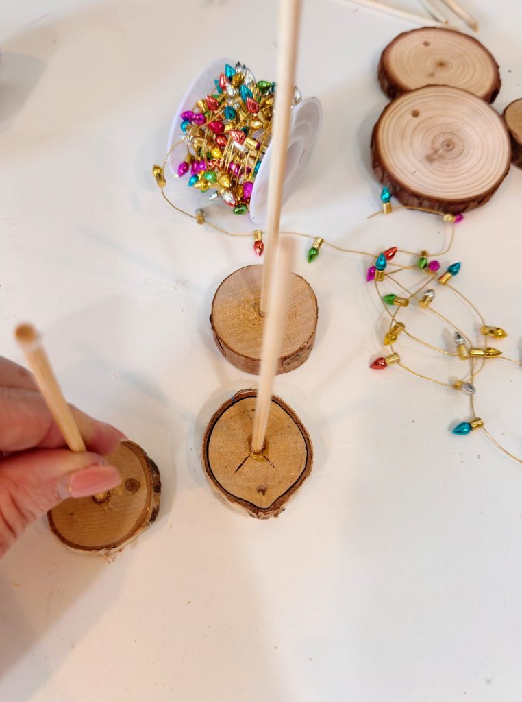 A hand inserts a wooden stick into a round wooden base. More wooden bases, sticks, and colorful decorative string lights are on the table.