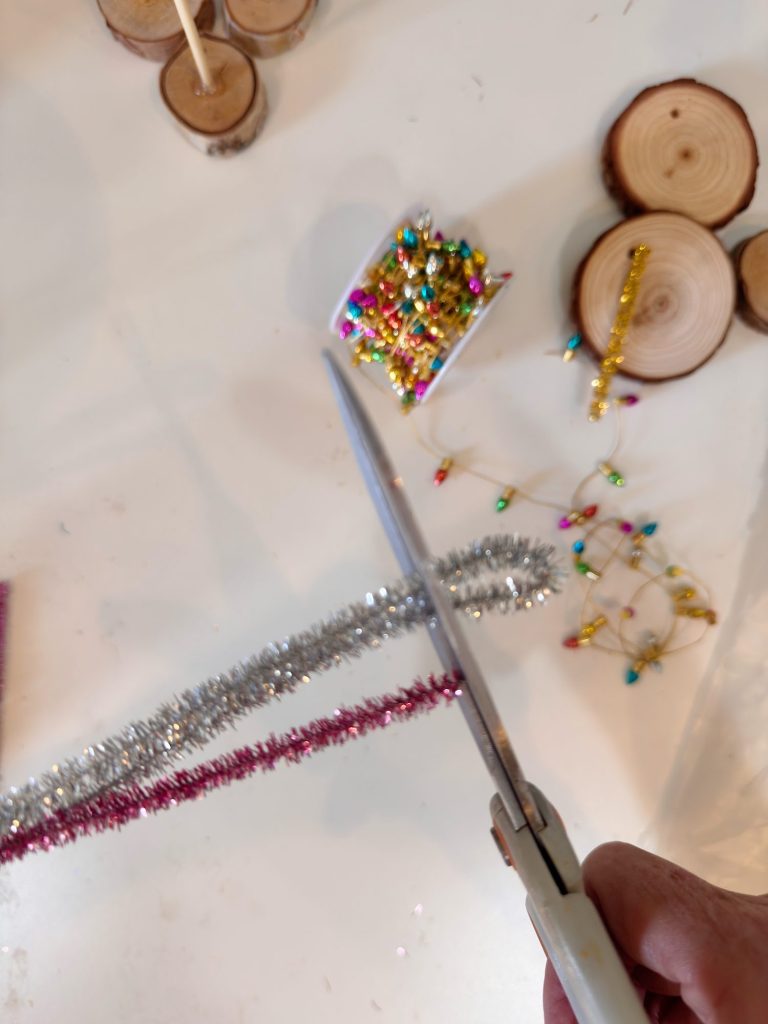 A hand holds scissors, cutting shiny pipe cleaners beside wooden slices and colorful beads on a white surface.
