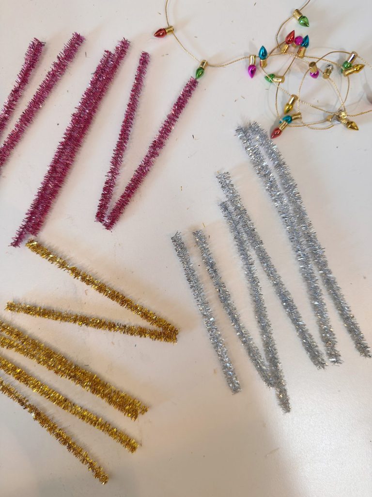 Three groups of metallic pipe cleaners in red, gold, and silver are arranged on a white surface next to a small string of multicolored holiday lights.