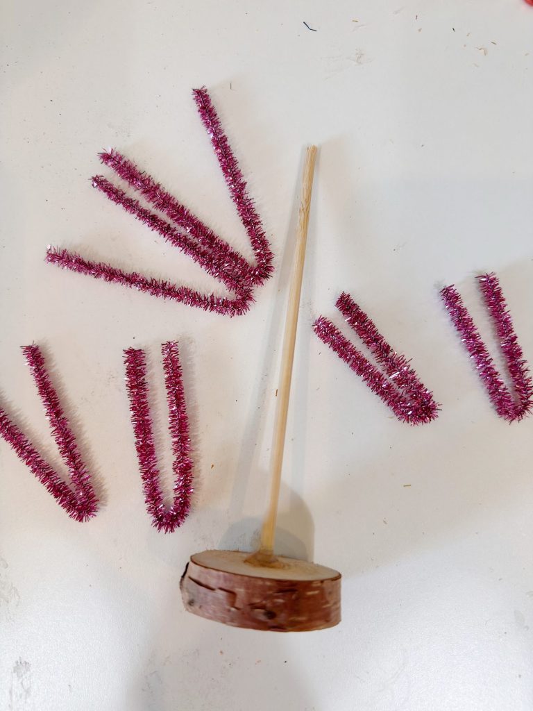 A wooden base with a vertical stick is surrounded by six pieces of red tinsel pipe cleaners, bent in various shapes, on a white surface.