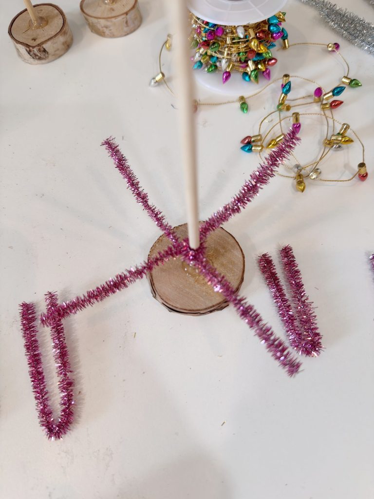 A craft project with a wooden disc base, pink pipe cleaners arranged in an X shape, a vertical stick, and holiday light garlands in the background on a white surface.