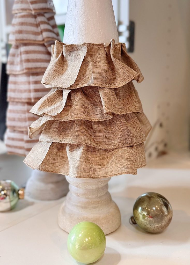 A white cone-shaped decoration with layered beige fabric ruffles stands on a white surface beside two round ornaments, one green and one metallic.