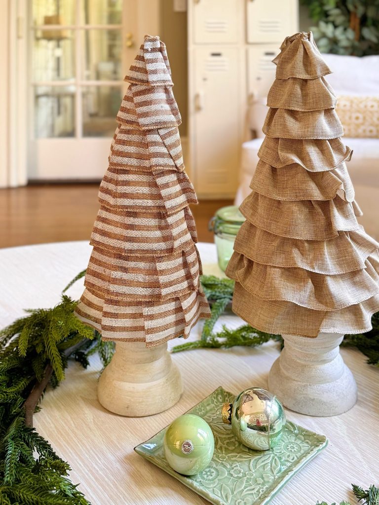 Two decorative fabric Christmas trees on white stands sit on a table beside green garland and three ornaments on a green tray in a cozy, well-lit room.