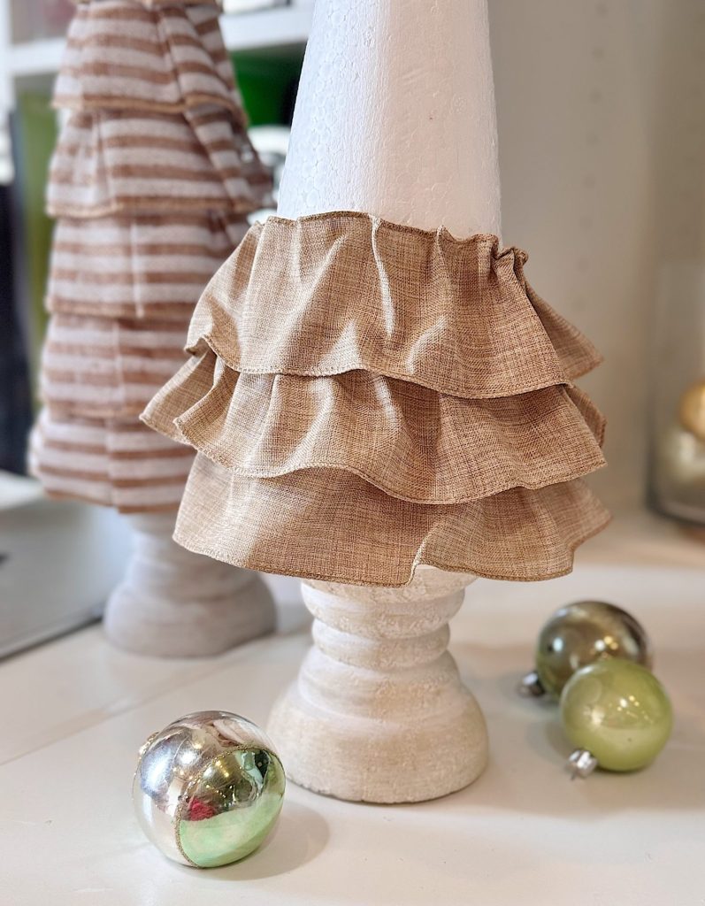 A white cone-shaped decoration with two layers of beige ruffled fabric is displayed on a white surface next to silver and green ornaments.