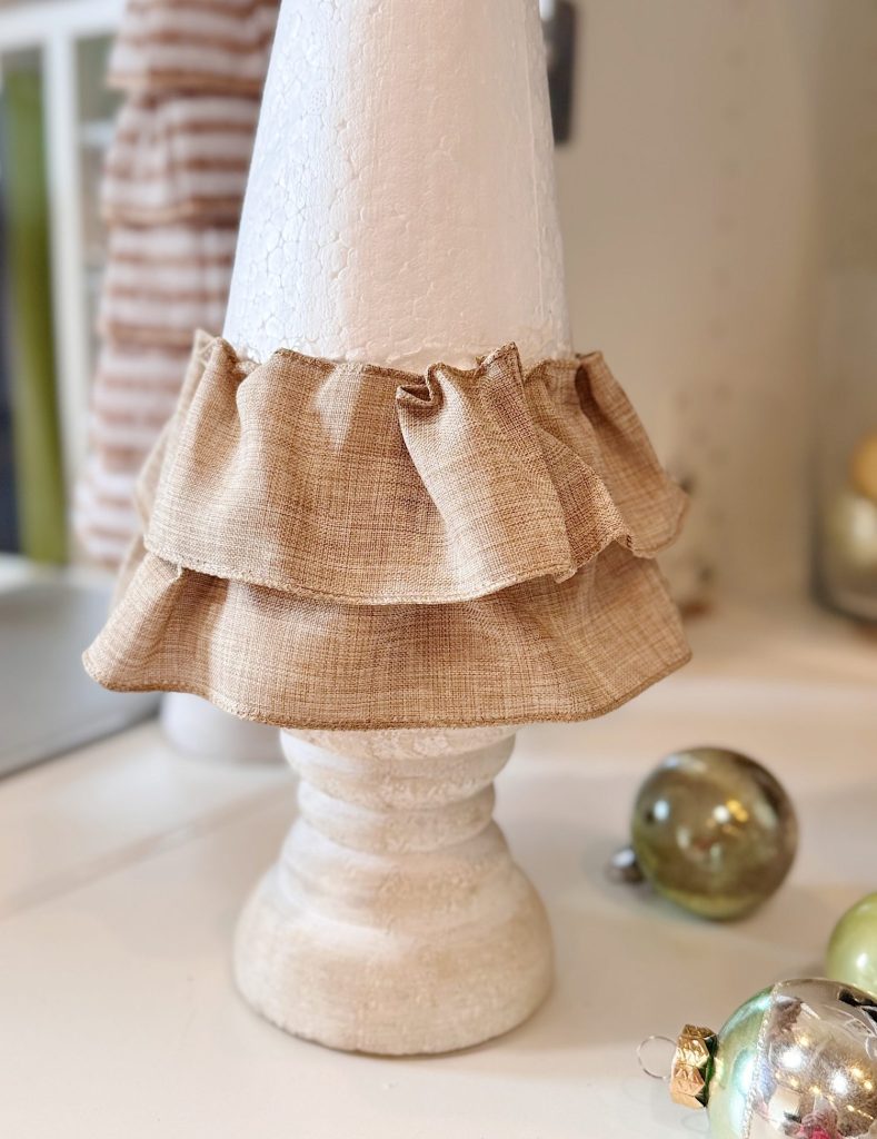 A white cone-shaped object with two layers of beige ruffled fabric around it, placed on a white surface with ornaments nearby.